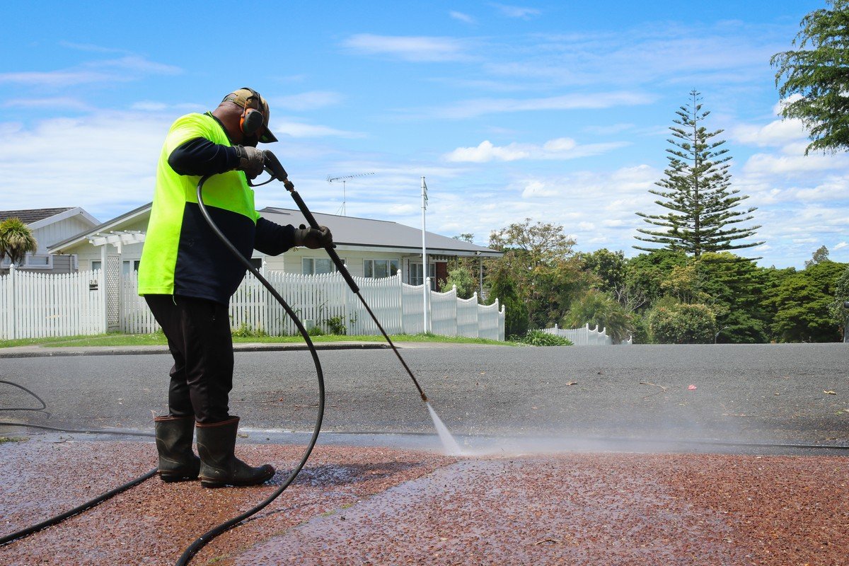 Worker in high-vis gear pressure washing a residential driveway with stream of water visible against red aggregate surface.