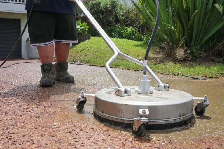 Worker operating a large rotary surface cleaner on wet red brick driveway with residential home and garden visible.