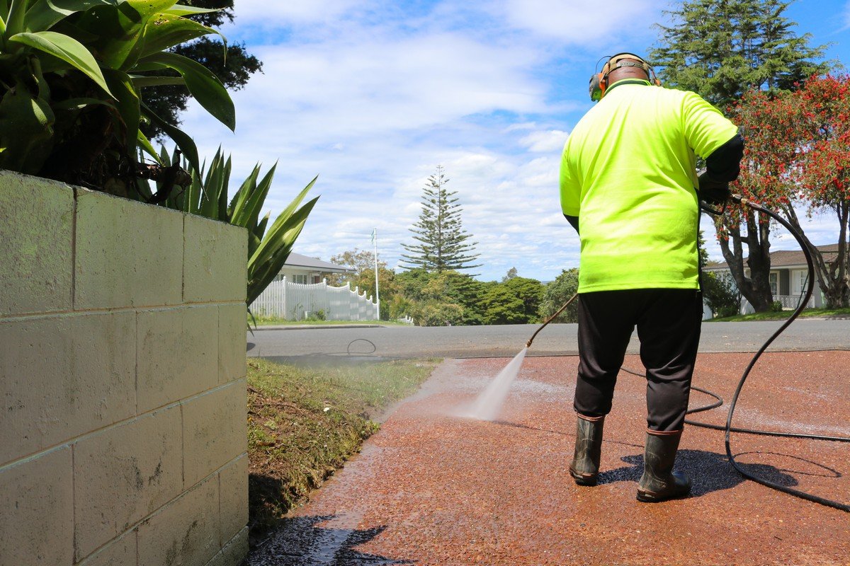 Worker in high-visibility jacket pressure washing a red brick driveway with concrete wall and landscaping visible.