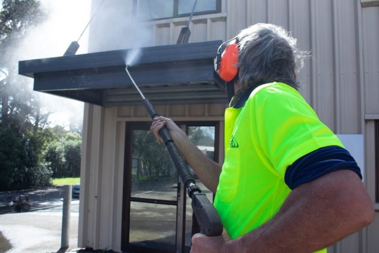 Worker in fluorescent vest with hearing protection pressure washing black awning and fascia of modern building entrance.